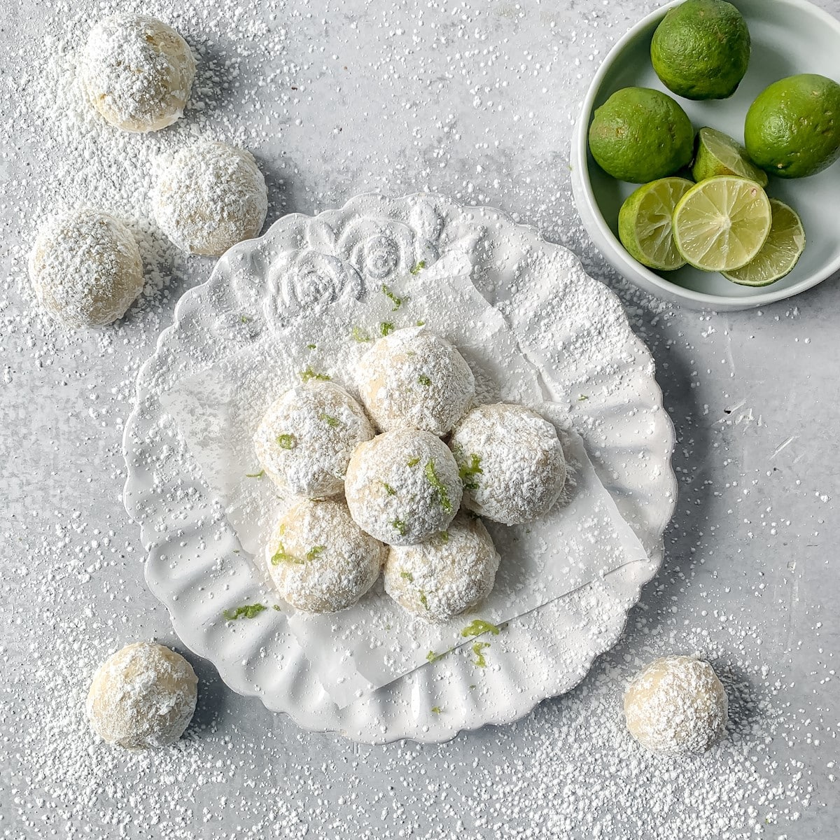 key lime cookies on a white plate, limes in a white bowl, cookies and powdered sugar on the side.