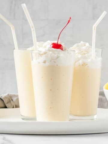 Three mango milkshakes in glasses with whipped cream, straws and a cherry.