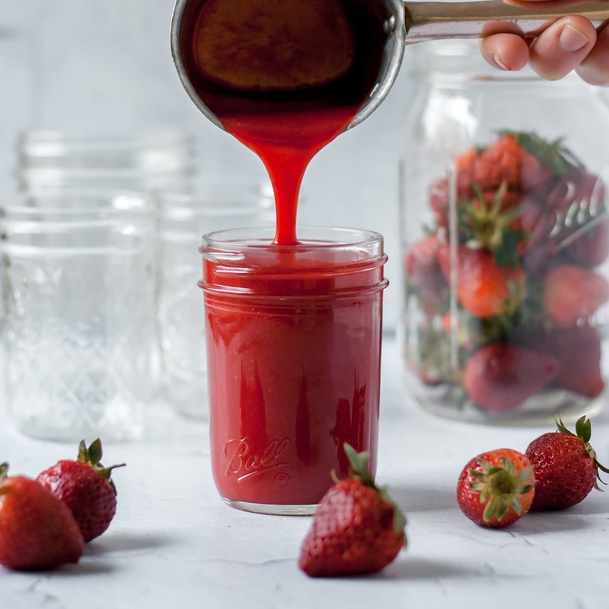 Strawberry coulis being poured into a mason jar, strawberries in background.