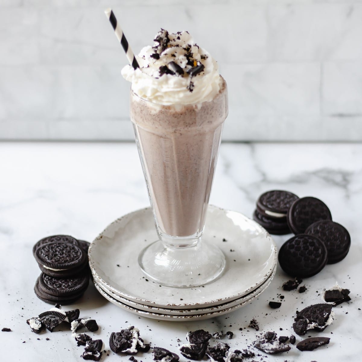 Cookies and cream milkshake in a glass, on a stack of plates with oreo cookies around it.
