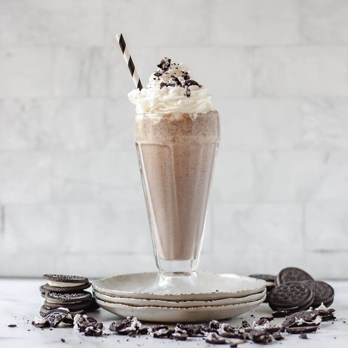 Cookies and cream milkshake in a glass, on a stack of plates with oreo cookies around it.