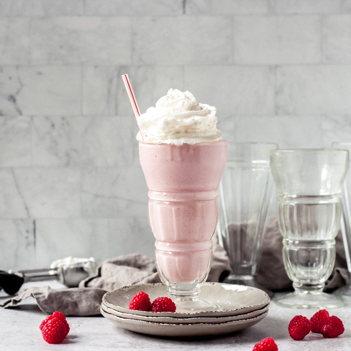 Raspberry milkshake in a glass on stacked plates with fresh raspberries.